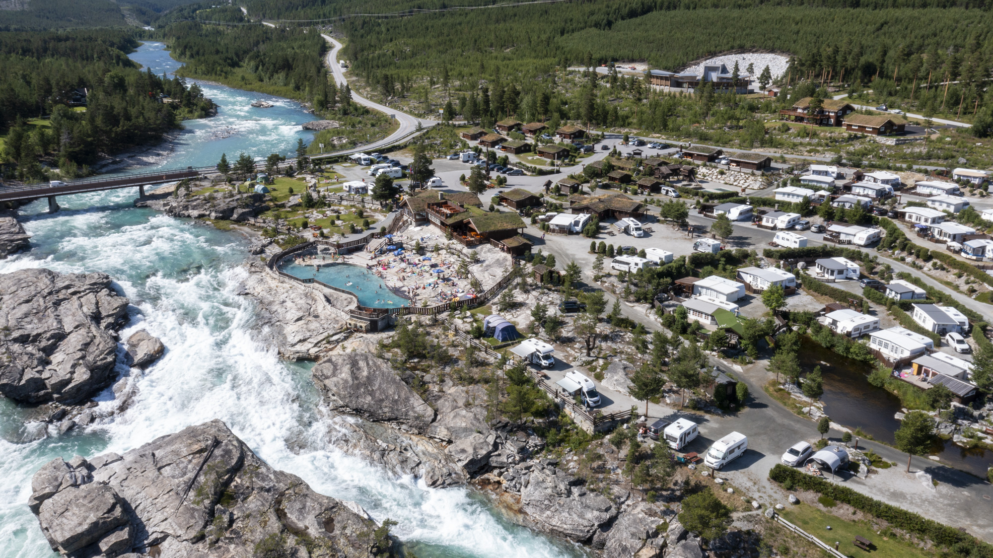 Oversiktsbilde av Dønfoss camping. Fossen renner forbi, folk bader og restauranten er åpen.