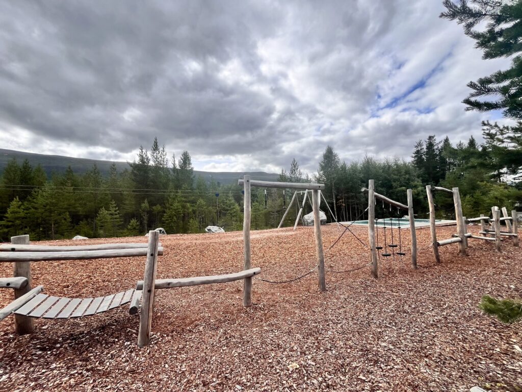Klatrestativ med balansebom og tau på lekeplassen, med trampoliner og fjell synlig i bakgrunnen.