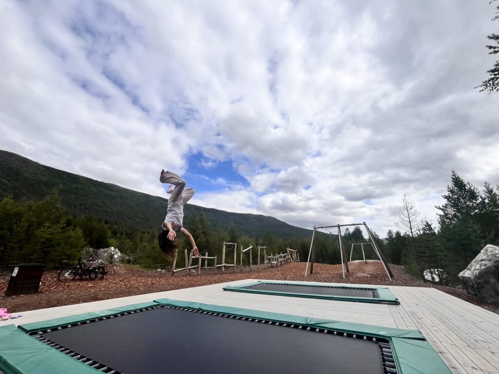 Barn tar salto over trampolinen, med klatrestativ og sykkel synlig på lekeplassen i bakgrunnen.