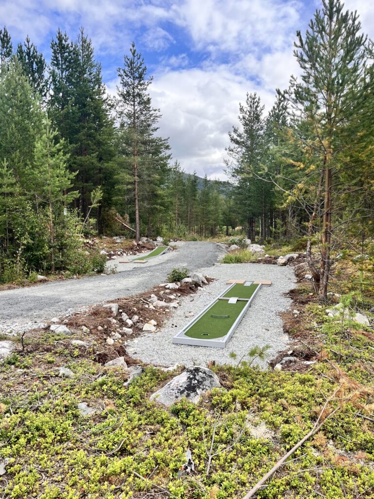 Minigolfbane med kunstgresshull langs grusstien i furuskogen, med fjell i det fjerne.