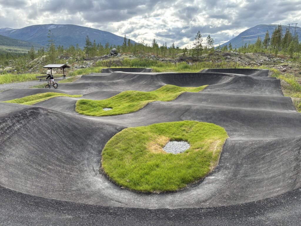 Asfaltert pumptrack med grønne gressøyer, syklist i bakgrunnen og fjell i horisonten.