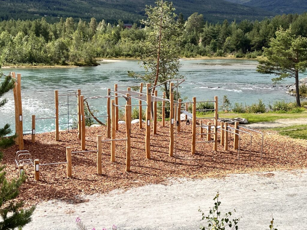 Utendørs treningsapparater i tre og stål ved elvebredden, med fjell i bakgrunnen.